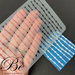 May include: A clear plastic stencil with a repeating pattern of lines and circles. The stencil is being held over a blue surface with a white pattern of lines and circles.