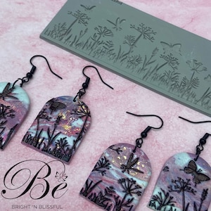 May include: A pair of handmade polymer clay earrings with a whimsical nature theme. The earrings feature a pink and white marbled background with gold accents and black silhouette images of wildflowers, butterflies, and dragonflies. The earrings are shaped like an arch and hang from black hooks.
