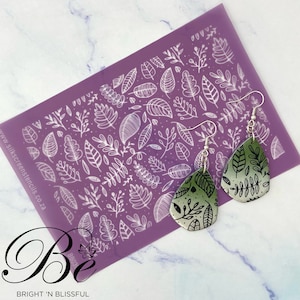 May include: A pair of green and silver earrings with a leaf pattern. The earrings are on a purple background with a white leaf pattern.