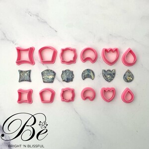 May include: A set of 12 pink plastic cookie cutters and 12 matching resin cabochons with a marbled design. The cutters are shaped like a square, a rounded rectangle, a rectangle with rounded corners, a crescent moon, a tulip, and a teardrop. The cabochons are the same shapes as the cutters.