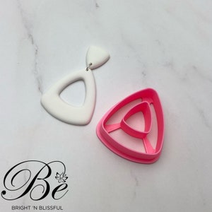 Könnte beinhalten: Ein weißer, dreieckiger Ohrring mit einem kleineren dreieckigen Ausschnitt in der Mitte. Daneben befindet sich eine rosa Plastikform zum Herstellen des Ohrrings.