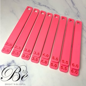 Puede incluir: Un conjunto de ocho reglas de medida de plástico rosa con los números 2.0, 2.5, 3.0, 3.5, 4.0, 5.0, 5.5 y 6.0 impresos en ellas.