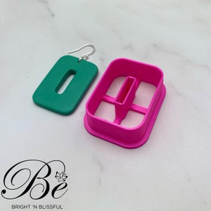 Può includere: Un orecchino rettangolare verde con un ritaglio a forma di rettangolo. L'orecchino è mostrato accanto a uno stampo in plastica rosa per la realizzazione dell'orecchino.