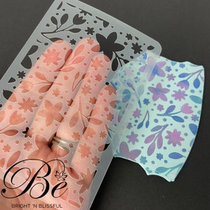 Puede incluir: Una plantilla de plástico transparente con un diseño floral. La plantilla se está utilizando para crear un patrón similar en una tela azul con flores moradas.