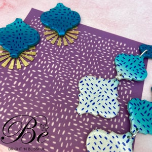 Puede incluir: Un par de pendientes de cerámica con estampado azul verdoso y blanco con detalles dorados. Los pendientes tienen forma de trébol de cuatro hojas y un degradado de color de oscuro a claro. Los pendientes están sobre un fondo morado con un patrón de lunares blancos.