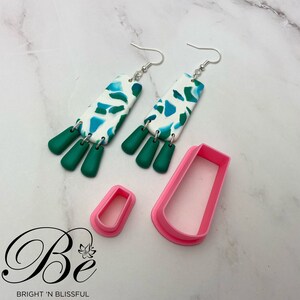 Pode incluir: Um par de brincos pendentes com um padrão abstrato branco e verde, e dois cortadores de argila polimérica rosa. Os brincos têm ganchos prateados e pingentes em forma de lágrima verde. Os cortadores têm a forma de um retângulo e uma lágrima.