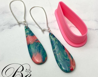 May include: Teardrop earrings with a marbled effect in teal, coral, and gold, alongside a pink teardrop-shaped cutter. The earrings feature silver-coloured ear wires. The logo "Be BRIGHT 'N BLISSFUL" is visible in the lower left corner.