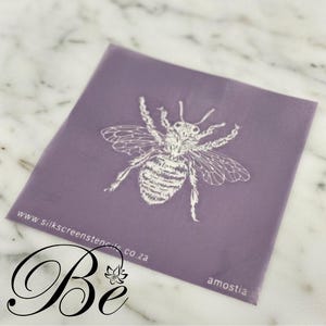 Peut inclure: Un pochoir sérigraphique lavande avec une illustration blanche d'abeille. Le pochoir comprend le texte "www.silkscreenstencils.co.za" et le mot "amostia". Le mot "Be" est écrit en cursive, avec un motif floral.