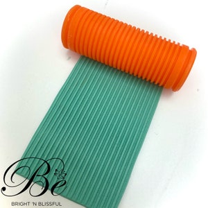 May include: An orange ribbed rolling pin is pressing into a teal, textured surface. The surface has parallel lines. The image has the text "Be" and "BRIGHT 'N BLISSFUL" in the bottom left corner.