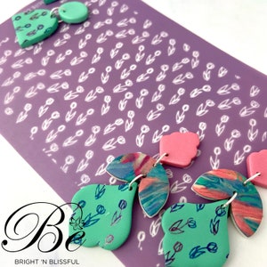 May include: A pair of handmade polymer clay earrings featuring a floral design. The earrings are made up of three pieces: a large teardrop shape, a smaller teardrop shape, and a small round shape. The earrings are a mix of green, pink, and blue colours. The earrings are shown on a purple background with a white tulip pattern.