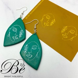 May include: A pair of green teardrop-shaped earrings with a white line drawing of a face and leaves. The earrings are on a white surface with a gold card behind them. The card has the text "Silk Screen Silks Co. 2A" printed on it.