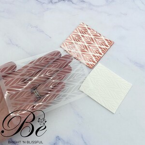May include: A clear stencil with a leaf pattern is held over a white surface. A copper-colored square with the same leaf design and a white square with an embossed leaf pattern are also visible. The logo "Be BRIGHT 'N BLISSFUL" is in the bottom left corner.