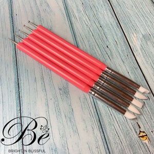 Può includere: Set di cinque strumenti per nail art con manico rosa e punte in silicone di diverse forme. Gli strumenti sono disposti in fila su una superficie in legno blu chiaro.
