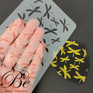 Peut inclure: Un pochoir en plastique transparent avec un motif de libellule. Le pochoir est placé sur une surface noire avec un motif de libellule dorée.