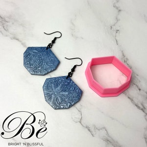 May include: A pair of blue and silver earrings with a floral design. The earrings are made of polymer clay and have a silver finish. The earrings are hanging from a black wire.