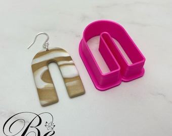 Könnte beinhalten: Ein Paar braune und weiße bogenförmige Ohrringe mit einem silbernen Haken. Die Ohrringe sind aus Polymer Clay gefertigt und haben einen marmorierten Effekt. Eine rosa Plastikform der Bogenform ist ebenfalls abgebildet.