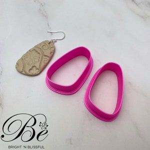 May include: Two pink plastic earring molds, one on top of the other, on a white marble surface. A beige clay earring with a floral design is in the background.