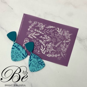 May include: Teal blue triangle shaped earrings with a floral pattern. The earrings are next to a purple stencil with a white floral design. The stencil has the text "www.silkscreenstencils.co.za".