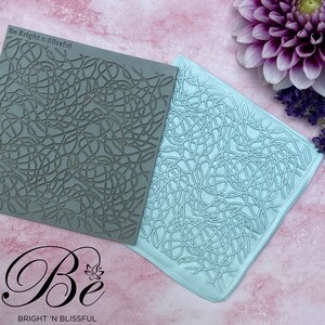 Könnte beinhalten: Zwei Silikoneinprägematten mit einem verworrenen Linienmuster. Die Matten sind grau und hellblau. Die Matten liegen auf einer rosa Oberfläche. Der Text "Be Bright 'n Blissful" befindet sich auf der grauen Matte.