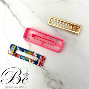 Może przedstawiać: Trzy spinki do włosów, jedna jest złota, jedna różowa, a jedna kolorowa z abstrakcyjnymi wzorami. Spinki są prostokątne i mają prosty design.