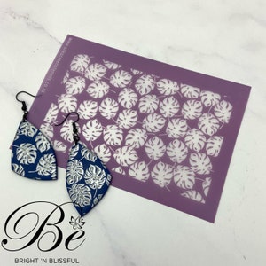 May include: Two blue and silver earrings with a monstera leaf pattern, next to a purple stencil with a white monstera leaf pattern. The stencil is labelled "www.silkscreenstencils.co.za".