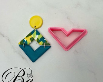 May include: A pair of geometric earrings with a teal, yellow and green pattern. The earrings are made of polymer clay and are shaped like a diamond. The earrings are shown next to a pink plastic heart-shaped cookie cutter.
