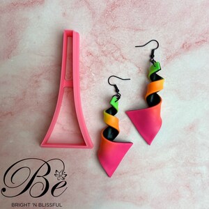 May include: A pair of pink, orange, green and black swirl earrings with black hooks. The earrings are on a pink marble surface with a pink plastic stand.