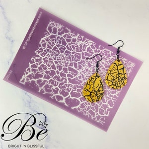 May include: A pair of yellow and black crackle effect teardrop earrings with black hooks. The earrings are displayed on a purple stencil with a white crackle pattern. The stencil has the text "www.silkscreenstencils.co.za" and "PS190C" printed on it.