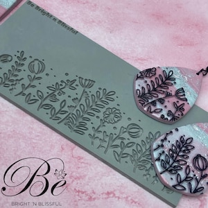 Op de afbeelding: Een grijze siliconen mal met een bloemenpatroon. De mal wordt gebruikt om oorbellen van polymer clay te maken. De oorbellen zijn roze en wit met zwarte bloemdetails. De tekst "Be Bright 'n Blissful" is op de mal gedrukt.