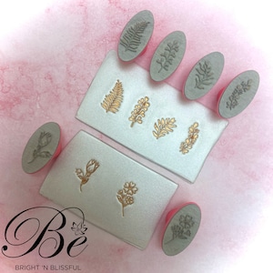 Puede incluir: Conjunto de nueve sellos de goma de forma ovalada con diseños florales y de hojas. Los sellos están montados en mangos de plástico rosa. Los sellos están dispuestos en dos superficies blancas con el texto "Be Bright 'N Blissful" impreso en una superficie.