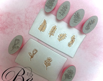 Puede incluir: Conjunto de nueve sellos de goma de forma ovalada con diseños florales y de hojas. Los sellos están montados en mangos de plástico rosa. Los sellos están dispuestos en dos superficies blancas con el texto "Be Bright 'N Blissful" impreso en una superficie.