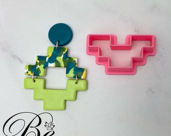 May include: A pink plastic cookie cutter shaped like a pixelated heart, next to a green, blue, and white geometric polymer clay earring. The earring is attached to a teal blue round polymer clay charm.