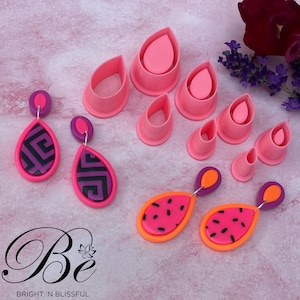 Könnte beinhalten: Ein Set aus rosa Polymer Clay-Ausstechern in verschiedenen Tropfenformen sowie zwei Paar handgefertigte Polymer Clay-Ohrringe. Ein Paar zeigt ein rosa-schwarzes geometrisches Muster, das andere Paar zeigt ein rosa-schwarzes Wassermelonen-Design. Die Ohrringe hängen an einer rosa Oberfläche mit dem Text "Be Bright 'n Blissful".