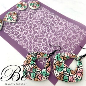 May include: A pair of handmade earrings featuring a geometric design in shades of pink, green, gold, and black. The earrings are crafted from polymer clay and have a unique, intricate pattern. The earrings are displayed against a purple background with a white stencil.