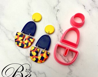May include: A set of two pink polymer clay cutters for making earrings. The cutters are shaped like a half-circle and a rectangle with a rounded top. The cutters are shown next to a pair of earrings made with the cutters. The earrings are made of blue, yellow, and orange polymer clay. The earrings are shaped like a half-circle and a rectangle with a rounded top. The earrings are hanging from a silver hook.