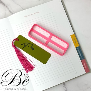 May include: A green bookmark with a pink tassel and a black line drawing of a person doing yoga. The bookmark is lying on a white page with lined paper. The page is part of a notebook with pink, yellow, and orange tabs that say "You Time", "Something New", and "Goals". The notebook also has the text "Be Real. Be Free. Be Happy." and "Be BRIGHT 'N BLISSFUL" printed on the page.