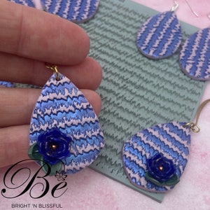 May include: A pair of blue and white striped teardrop-shaped earrings with a blue flower accent. The earrings are made of polymer clay and have a gold hook.