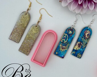 Può includere: Due paia di orecchini in resina con dettagli dorati. Il primo paio è di colore grigio chiaro con glitter dorati e il secondo paio è di colore blu con dettagli dorati. Gli orecchini sono mostrati con uno stampo in silicone rosa.
