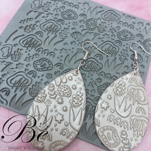 May include: A pair of silver teardrop-shaped earrings with a white background and a raised design of abstract shapes and flowers. The earrings are shown on a gray silicone sheet with a similar design.