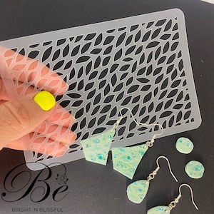 May include: A clear plastic stencil with a repeating pattern of leaf shapes. The stencil is being used to create earrings with a green and blue glitter finish. The earrings are in the shape of a triangle, a teardrop, and a circle.