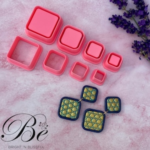 Può includere: Un set di otto tagliapasta quadrati in plastica rosa di diverse dimensioni. I tagliapasta sono disposti in uno schema sfalsato su una superficie rosa. Sono mostrati anche due paia di orecchini quadrati blu e dorati.