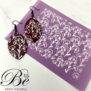 May include: A pair of burgundy and white floral earrings with a matching stencil. The stencil is purple and features a repeating pattern of white flowers.