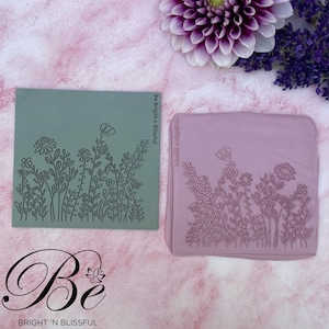 May include: Two square, floral-themed stamps. One is sage green, the other is mauve. Each features a detailed outline of wildflowers and foliage. The stamps are set against a pink background with a dahlia and lavender sprig. The text "Be Bright 'n Blissful" is visible.