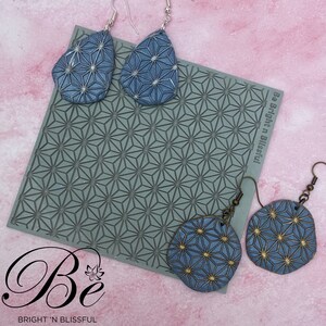 May include: A set of four blue and gold earrings with a geometric pattern. The earrings are made of polymer clay and are hanging from silver ear wires. The earrings are displayed on a green silicone mat with a geometric pattern. The mat has the text "Be Bright 'n Blissful" printed on it.