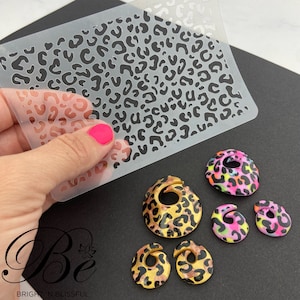 May include: A set of five leopard print polymer clay earrings. The earrings are in various colors, including pink, black, gold, and white. The earrings are shown on a black surface with a clear plastic stencil of a leopard print pattern.