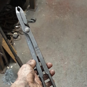 Może przedstawiać: Szczypce kowalskie z długimi uchwytami i chwytającą głowicą. Metalowe narzędzie trzymane jest w dłoni, ukazując otoczenie warsztatu. Szczypce mają wytłoczone litery "SLB". Narzędzie służy do trzymania gorącego metalu.