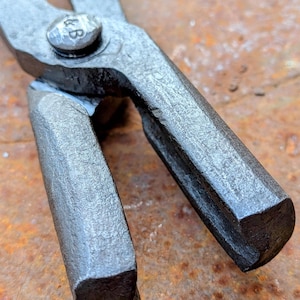 Może przedstawiać: Zbliżenie pary metalowych szczypiec, prawdopodobnie do kowalstwa. Szczypce są w ciemnoszarym kolorze, z teksturowaną, lekko szorstką powierzchnią. Na nicie łączącym dwa ramiona widnieją inicjały "&B". Tło jest w kolorze rdzy, pomarańczowo-brązowym.