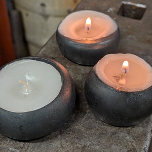 Könnte beinhalten: Drei brennende Kerzen in dunkelgrauen, abgerundeten Haltern. Die Kerzen haben einen weißen Wachsinnenraum und ein warmes, orangefarbenes Leuchten von den Flammen. Die Halter stehen auf einer strukturierten, grauen Oberfläche.