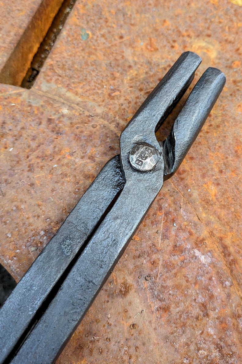 Może przedstawiać: Zbliżenie czarnych metalowych szczypiec kowalskich z nitą w punkcie obrotu. Narzędzie ma teksturowaną powierzchnię i jest umieszczone na tle zardzewiałego, pomarańczowo-brązowego metalu. Na nicie wytłoczono litery "B&B".