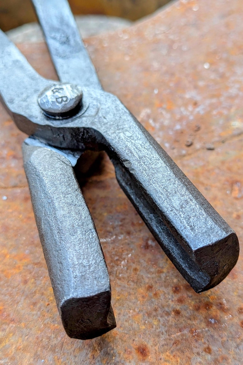 Może przedstawiać: Zbliżenie pary metalowych szczypiec, prawdopodobnie do kowalstwa. Szczypce są w ciemnoszarym kolorze, z teksturowaną, lekko szorstką powierzchnią. Na nicie łączącym dwa ramiona widnieją inicjały "&B". Tło jest w kolorze rdzy, pomarańczowo-brązowym.